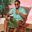 Load image into Gallery viewer, Seated view of happy view of male model wearing the adult 7-Strong &quot;Sunset Palms&quot; shirt, featuring small yellow and pink suns, reminiscent of a sunset, behind a silhouette of a palm tree and birds, patterned at various angles all over the turquoise-like shirt. The model is featured against a pink tropical backdrop with palm trees in the background.

