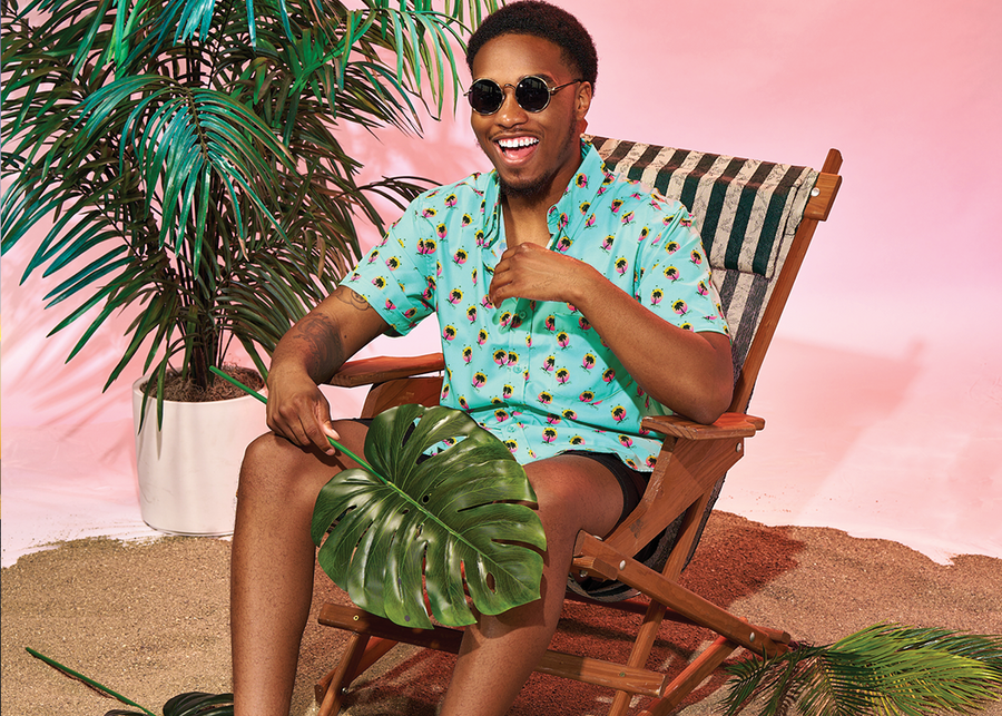 Seated view of happy view of male model wearing the adult 7-Strong "Sunset Palms" shirt, featuring small yellow and pink suns, reminiscent of a sunset, behind a silhouette of a palm tree and birds, patterned at various angles all over the turquoise-like shirt. The model is featured against a pink tropical backdrop with palm trees in the background.