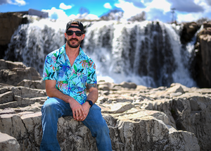 Male model, staged on the right of camera, sitting on rocks with a waterfall backdrop wearing the 7-Strong adult "Tropic of Conversation" button down - featuring various exotic rainforest flowers overlapping one another in a vibrant display against a white ghost background of other fronds and plants. 