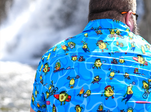 Back view of male model, facing away from camera, wearing the 7-Strong Avocado Coast button down featuring various avocado characters engaging in summer activities such as grilling, playing volleyball, weightlifting, surfing, etc against a blue shimmery wave looking background. 