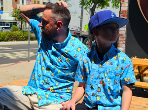 Male adult and male youth models wearing the 7-Strong Avocado Coast button down, sitting on a bench and looking in opposite directions. 