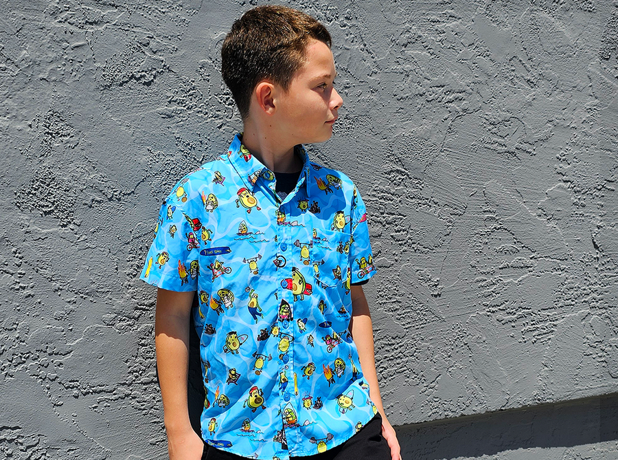 Youth male model standing against a gray stone wall, looking away from camera wearing the 7-Strong youth Avocado Coast button down. 