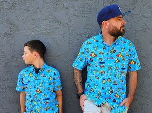 Male youth and male adult models wearing the 7-Strong Avocado Coast button down, against a gray stone wall looking away from camera and in opposite directions.  