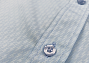 Close up mid button view of the baby blue long sleeve EveryWEAR adult button up. Pattern is subtle white 7-Bolts lined up all against each other across a baby blue background. The shirt is featured on a soft background of a wave hitting the beach. 