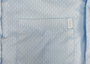Close up pocket view of the baby blue long sleeve EveryWEAR adult button up. Pattern is subtle white 7-Bolts lined up all against each other across a baby blue background. The shirt is featured on a soft background of a wave hitting the beach. 