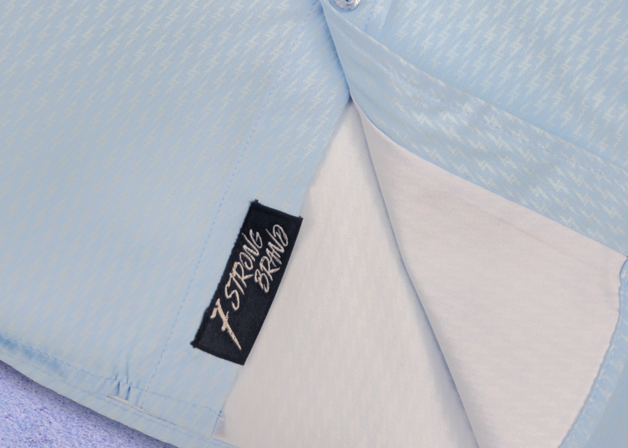 Bottom sweep tag view of the baby blue long sleeve EveryWEAR adult button up. Pattern is subtle white 7-Bolts lined up all against each other across a baby blue background. The shirt is featured on a soft background of a wave hitting the beach.