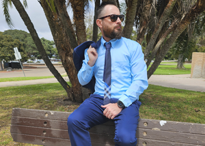 Shot of a male model, on a bench looking off into the distance in a blue suit accented by the 7-Strong EveryWEAR collection baby blue long sleeve shirt.