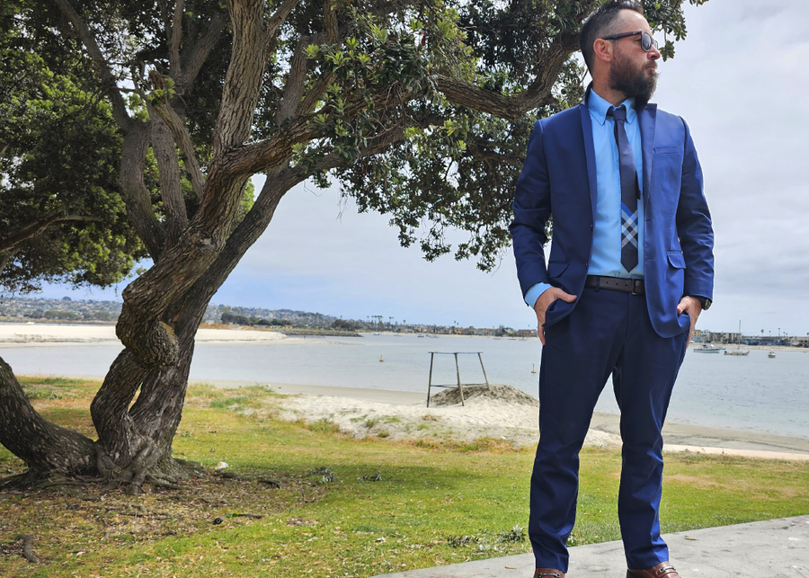 Shot of a male model, on a beach path looking off into the distance in a blue suit accented by the 7-Strong EveryWEAR collection baby blue long sleeve shirt. 