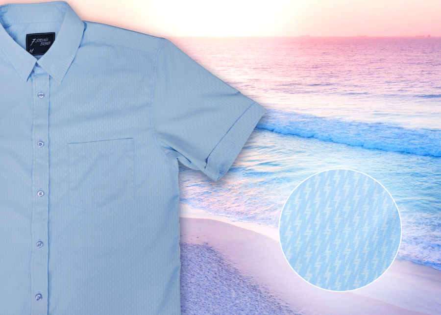 3/4 full view of the baby blue short sleeve EveryWEAR adult button up. Pattern is subtle white 7-Bolts lined up all against each other across a baby blue background. The shirt is featured on a soft background of a wave hitting the beach. Bottom right corner features a detail circle of the bolts up close. 