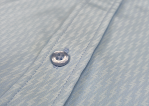 Close up mid button view of the baby blue short sleeve EveryWEAR adult button up. Pattern is subtle white 7-Bolts lined up all against each other across a baby blue background. The shirt is featured on a soft background of a wave hitting the beach. 