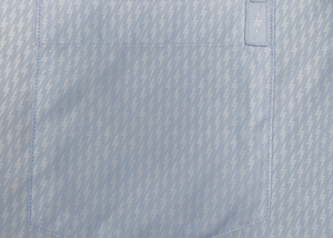 Close up of the seamless pocket view, with bolt pocket tag, of the baby blue short sleeve EveryWEAR adult button up. Pattern is subtle white 7-Bolts lined up all against each other across a baby blue background. The shirt is featured on a soft background of a wave hitting the beach.
