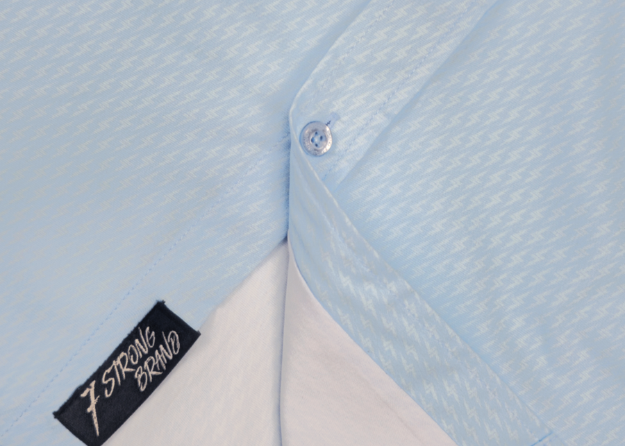 Bottom sweep tag view of the baby blue short sleeve EveryWEAR adult button up. Pattern is subtle white 7-Bolts lined up all against each other across a baby blue background. The shirt is featured on a soft background of a wave hitting the beach.