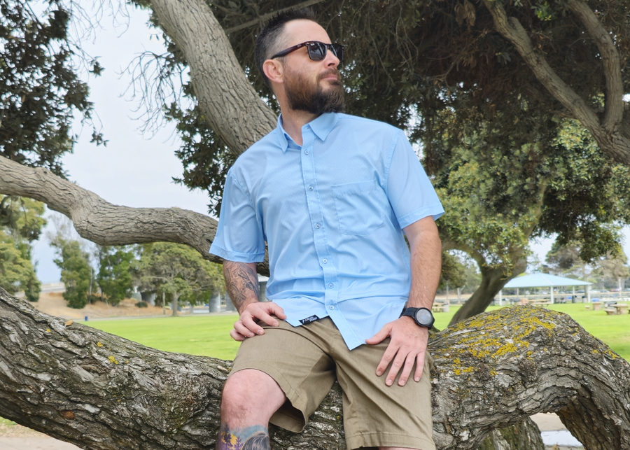 Medium shot of a male model, sitting on a log, looking off in to the distance wearing the baby blue 7-Strong EveryWEAR short sleeve button down. Design is baby blue with subtle white 7-bolts featured all over, side-by-side in white. 