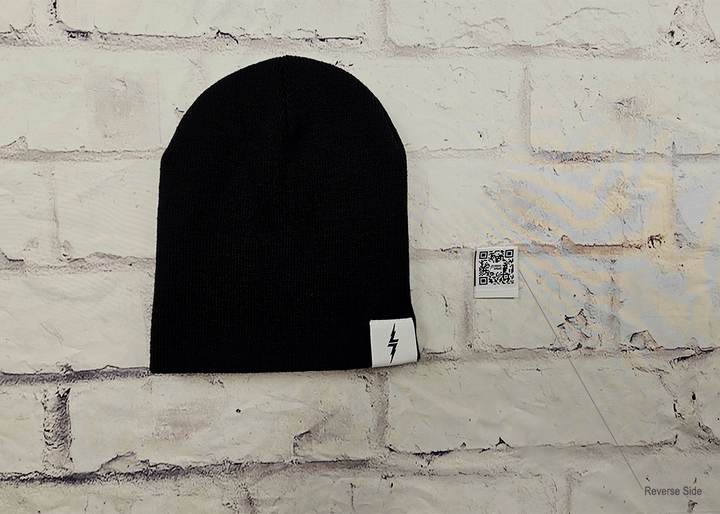 Full view of the black 7-Bolt beanie, rounded at the top and featuring a white 7-bolt tag in the bottom right corner of the cuff. Alongside is the reverse of the tag, featuring the 7-Strong QR Code. The beanie is featured against a white brick backdrop. 