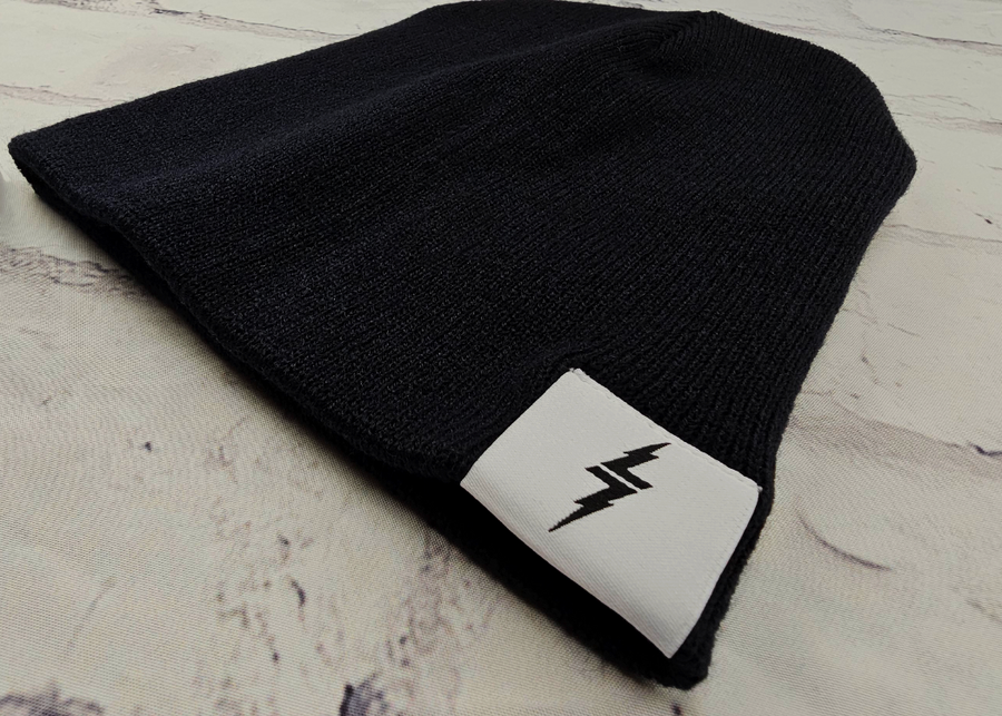 Angle full view of the black 7-Bolt beanie, rounded at the top and featuring a white 7-bolt tag in the bottom right corner of the cuff.  The beanie is featured against a white brick backdrop.