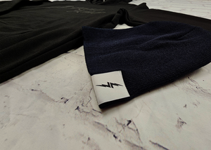 Angled view of the black 7-Bolt beanie, rounded at the top and featuring a white 7-bolt tag in the bottom right corner of the cuff. Alongside is the 7-Strong Midnight Hoodie. The beanie is featured against a white brick backdrop.