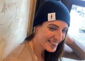 Female model smiling and looking away while wearing the black 7-Bolt beanie, reaching up to playfully adjust it.