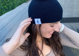 Female model smiling and looking away while wearing the black 7-Bolt beanie, reaching up to playfully adjust it. 