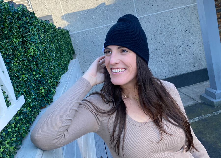 Female model smiling and looking up while wearing the black 7-Bolt beanie, reaching up to playfully adjust it.