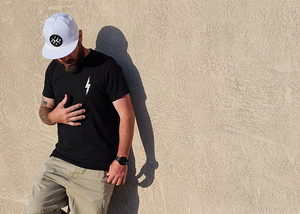 Male model, justified left of frame against a beige wall wearing the 7-Strong Black "Branded" Tee and white 7-Strong "Branded" white Performance hat, with raised X-Badge in black on the front. Model is looking down and away from camera. 