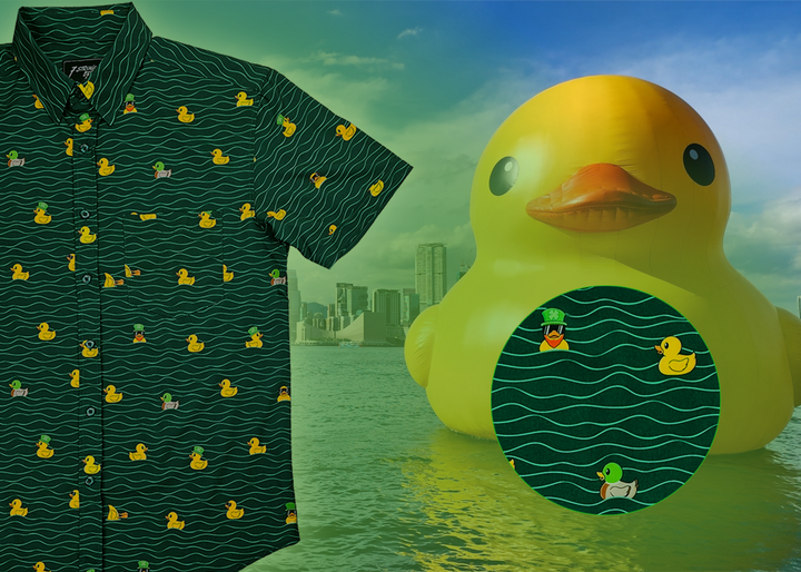 A 3/4 view of the 7-Strong "Duck O' The Irish" adult button down, featuring a deep green water-replicating background with various rubber ducks swimming along the waves. The shirt is featured against a green river scenic with a giant inflatable duck in the background. In the bottom right, there is a detail circle that shows the design pattern up close. 