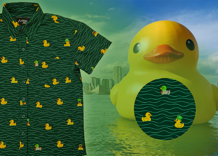 A 3/4 view of the 7-Strong "Duck O' The Irish" youth button down, featuring a deep green water-replicating background with various rubber ducks swimming along the waves. The shirt is featured against a green river scenic with a giant inflatable duck in the background. In the bottom right, there is a detail circle that shows the design pattern up close.