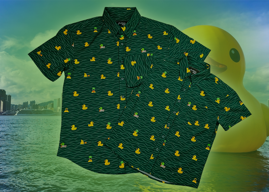 A centered, overlapping view of the 7-Strong "Duck O' The Irish" adult and youth button down, featuring a deep green water-replicating background with various rubber ducks swimming along the waves. The shirt is featured against a green river scenic with a giant inflatable duck in the background.
