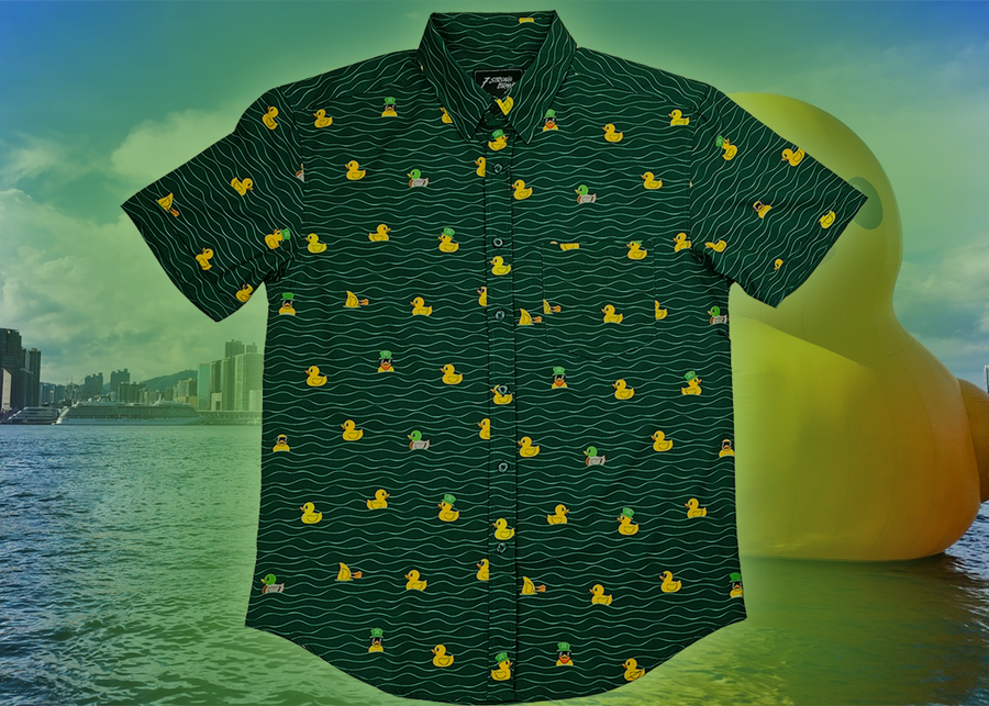 A centered view of the 7-Strong "Duck O' The Irish" adult button down, featuring a deep green water-replicating background with various rubber ducks swimming along the waves. The shirt is featured against a green river scenic with a giant inflatable duck in the background.