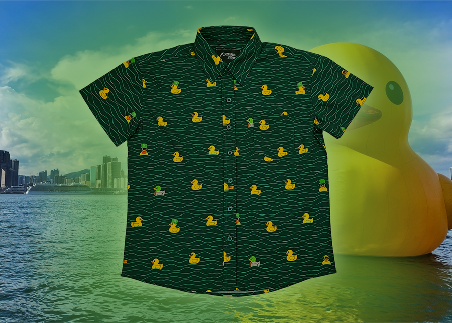 A centered view of the 7-Strong "Duck O' The Irish" youth button down, featuring a deep green water-replicating background with various rubber ducks swimming along the waves. The shirt is featured against a green river scenic with a giant inflatable duck in the background. 