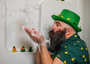 A male model sits in a bathtub wearing a green Irish top hat and blowing soap bubbles in his hand wearing the 7-Strong "Duck O' The Irish" adult button down, featuring a deep green water-replicating background with various rubber ducks swimming along the waves. 