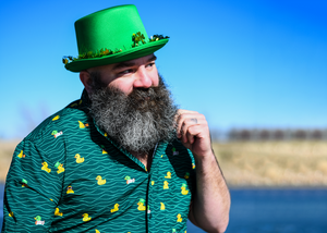 A male model standing by the water twirling his beard wearing a green Irish top hat and looking off while wearing the 7-Strong "Duck O' The Irish" adult button down, featuring a deep green water-replicating background with various rubber ducks swimming along the waves. 