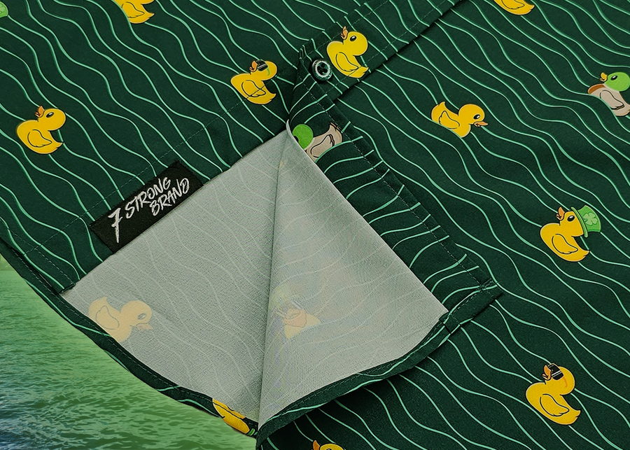A bottom sweep tag view of the 7-Strong "Duck O' The Irish" adult button down, featuring a deep green water-replicating background with various rubber ducks swimming along the waves. The shirt is featured against a green river scenic with a giant inflatable duck in the background.