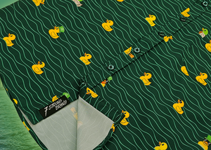 A bottom sweep tag view of the 7-Strong "Duck O' The Irish" youth button down, featuring a deep green water-replicating background with various rubber ducks swimming along the waves. The shirt is featured against a green river scenic with a giant inflatable duck in the background.