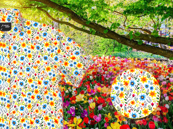 A 3/4 close-up shot of the 7-Strong "Daisy Me Rollin'" adult button down shirt, a white base color with various blue, red, orange, and yellow floral daisies in an all over print. The shirt is displayed against a background of a field of daisies. The bottom right features a detail circle with a close up of the shirt design. 