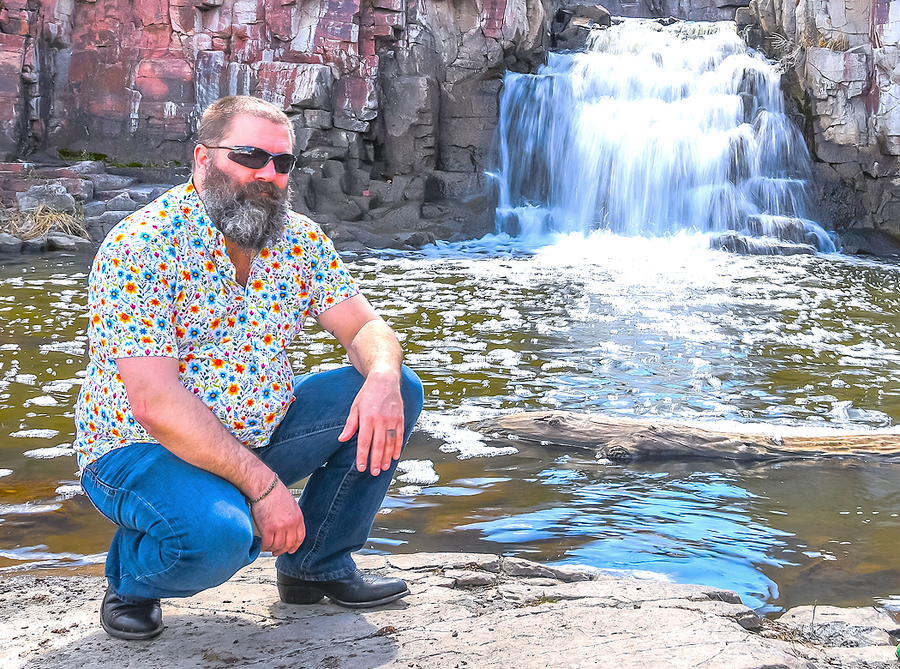 Photo of a a male model, squatting down facing camera, wearing the 7-Strong "Daisy Me Rollin'" adult button down shirt, a white base color with various blue, red, orange, and yellow floral daisies in an all over print. The model is out in front of a waterfall.