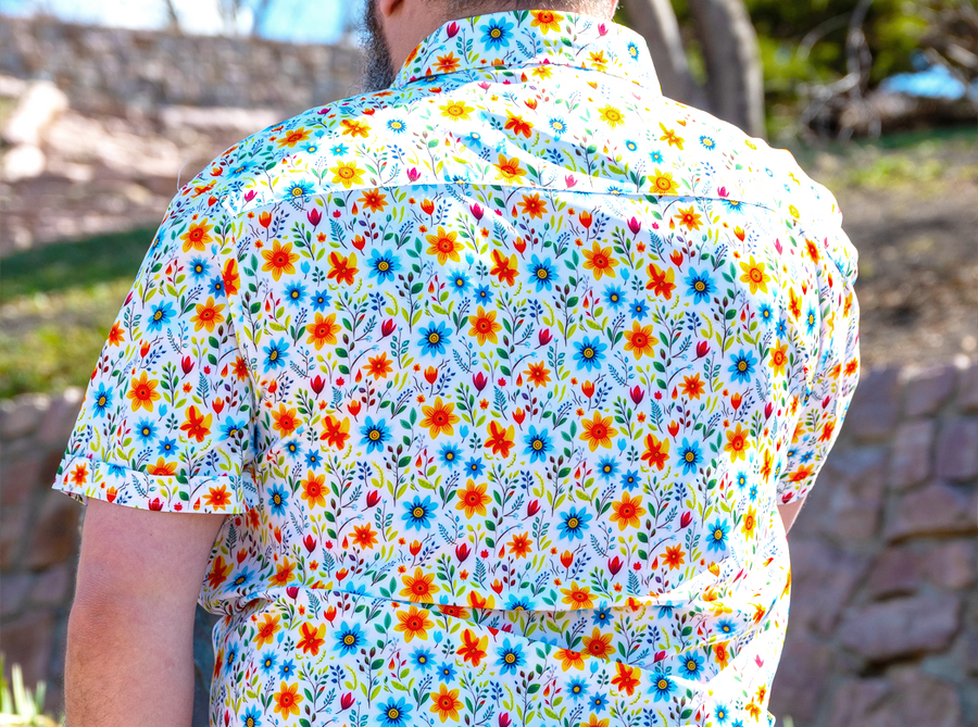 Behind the back look at a male model wearing the 7-Strong "Daisy Me Rollin'" adult button down shirt, a white base color with various blue, red, orange, and yellow floral daisies in an all over print. The model is facing away from camera standing out in nature.