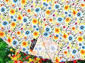 A bottom half close-up shot of the 7-Strong "Daisy Me Rollin'" adult button down shirt, a white base color with various blue, red, orange, and yellow floral daisies in an all over print. The shirt is displayed against a background of a field of daisies.