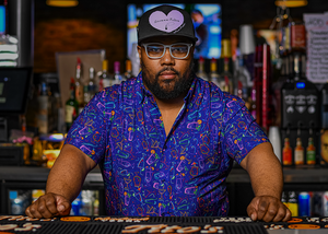 Male bartender standing behind the bar looking at the camera while wearing the 7-Strong "Happy Hour" button down shirt, featuring a multitude of neon colored drinks and glassware with garnishes, mixes, and the like all over a deep purple background.