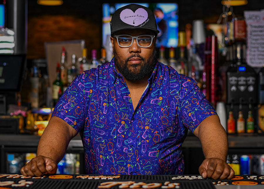 Male bartender standing behind the bar looking at the camera while wearing the 7-Strong "Happy Hour" button down shirt, featuring a multitude of neon colored drinks and glassware with garnishes, mixes, and the like all over a deep purple background.