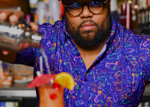 Male bartender crafting a mixed drink while wearing the 7-Strong "Happy Hour" button down shirt, featuring a multitude of neon colored drinks and glassware with garnishes, mixes, and the like all over a deep purple background. 