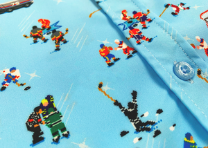 A midsection close up of the adult 8-Bit Hat Trick button down - featuring various 8-bit inspired scenes from hockey games on a light blue ice-like background. The shirt is featured against a ice hockey floor with hats strewn about. 