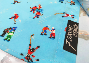 A bottom sweep tag view of the adult 8-Bit Hat Trick button down - featuring various 8-bit inspired scenes from hockey games on a light blue ice-like background. The shirt is featured against a ice hockey floor with hats strewn about. 
