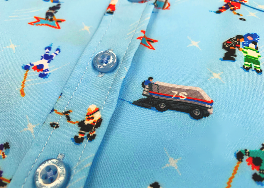A close up midsection view of the youth 8-Bit Hat Trick button down - featuring various 8-bit inspired scenes from hockey games on a light blue ice-like background. The shirt is featured against a ice hockey floor with hats strewn about.