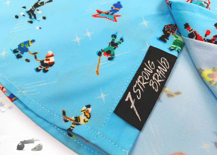 A bottom sweep tag view of the youth 8-Bit Hat Trick button down - featuring various 8-bit inspired scenes from hockey games on a light blue ice-like background. The shirt is featured against a ice hockey floor with hats strewn about.