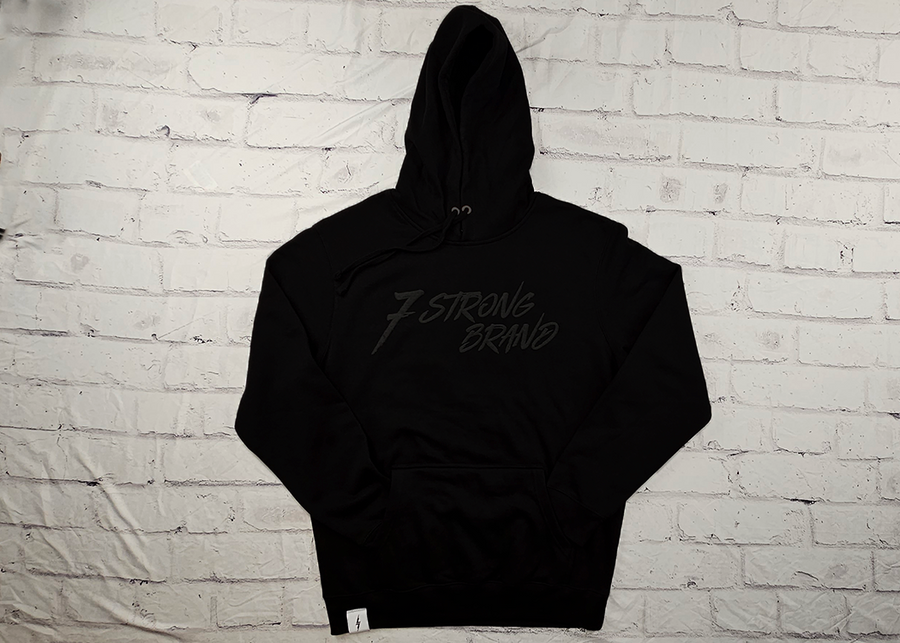 Full centered view of the 7-Strong Midnight Long Sleeve Hoodie, a black hoodie with black shadow font of the 7-Strong Brand logo. The hoodie is show against a white brick backdrop.
