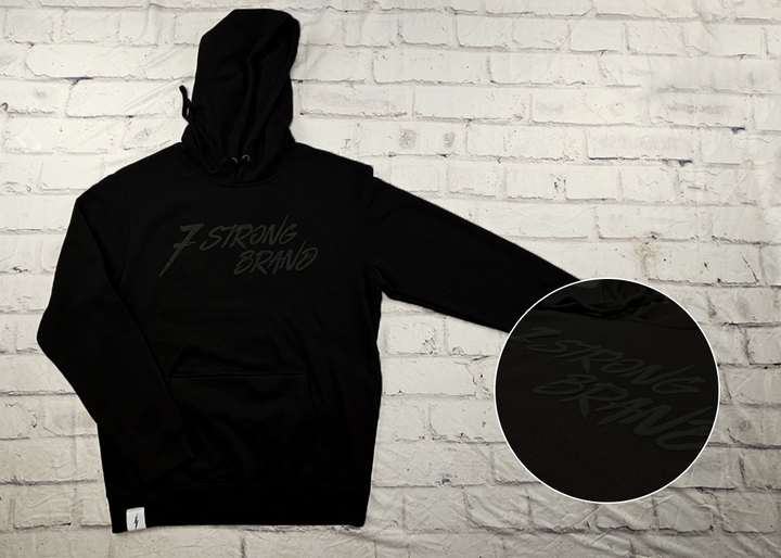 Full view of the 7-Strong Midnight Long Sleeve Hoodie, a black hoodie with black shadow font of the 7-Strong Brand logo. The hoodie is show against a white brick backdrop. The bottom right corner features a detail circle with the raised 7 Strong Brand logo across the chest. 