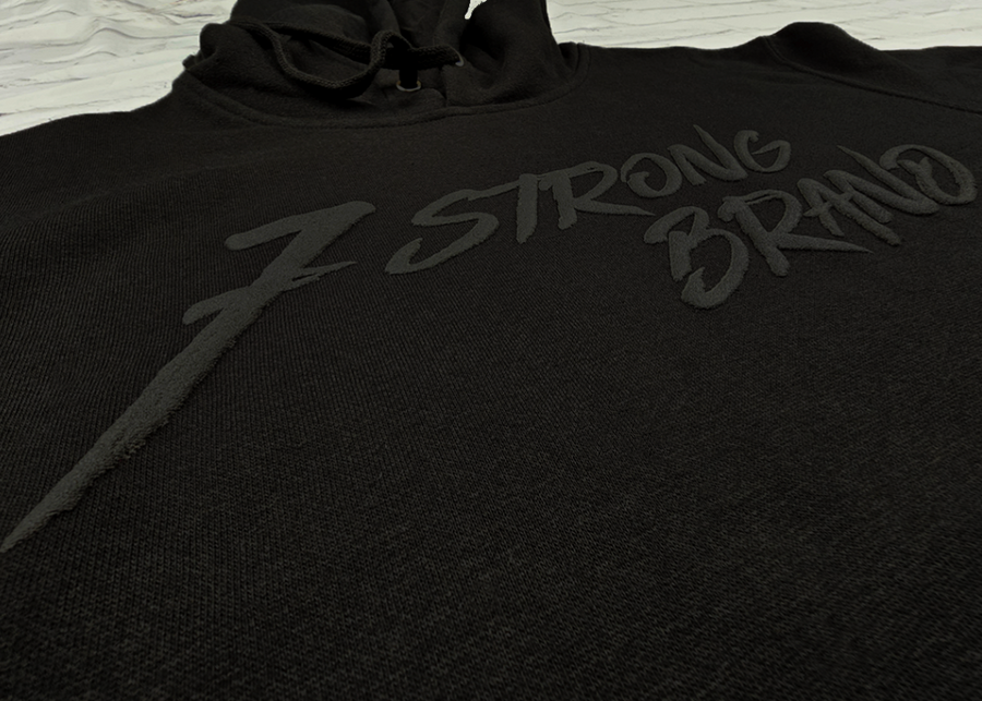 Midsection angled view of the 7-Strong Midnight Long Sleeve Hoodie, a black hoodie with black shadow font of the 7-Strong Brand logo. The hoodie is show against a white brick backdrop.