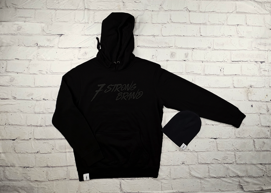 Full view of the 7-Strong Midnight Long Sleeve Hoodie, a black hoodie with black shadow font of the 7-Strong Brand logo. Alongside the black 7-Bolt Beanie with a white 7-Bolt tag on the cuff. The hoodie is show against a white brick backdrop.