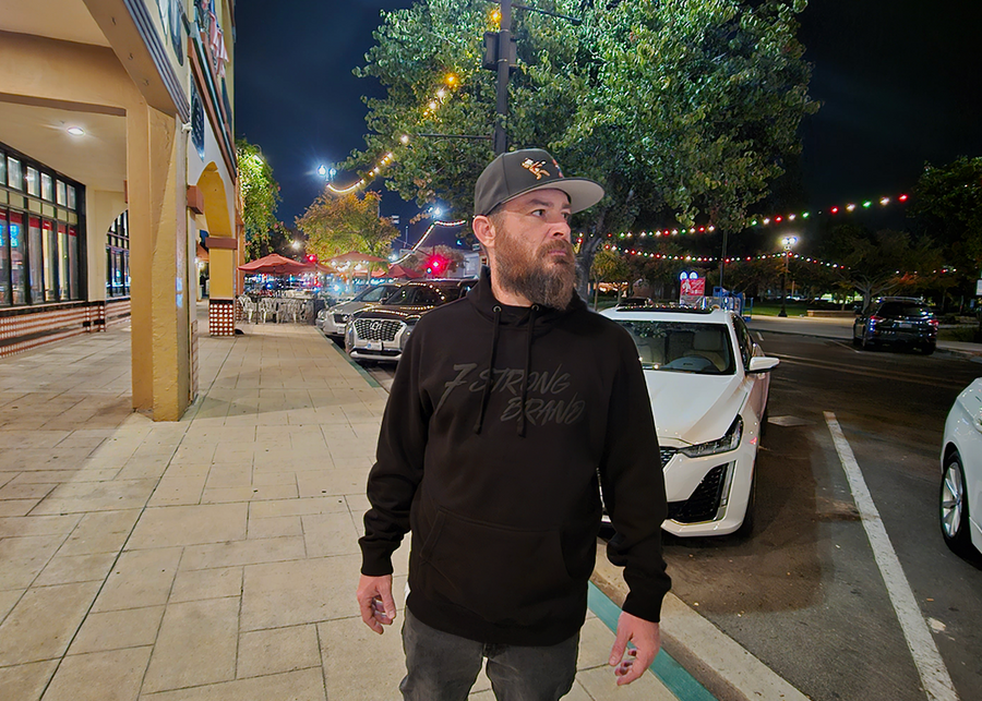 Male model out on the town looking off and wearing the 7-Strong Midnight Long Sleeve Hoodie, a black hoodie with black shadow font of the 7-Strong Brand logo. 