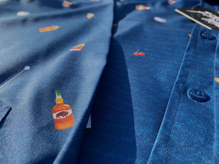 Midsection view of "Call Me Old Fashioned" adult long sleeve button-down. Features elements of an Old Fashioned cocktail (bottle of bourbon, glasses, oranges, stirrers, and ice) against a blue background which faintly spells out an Old Fashioned recipe. 